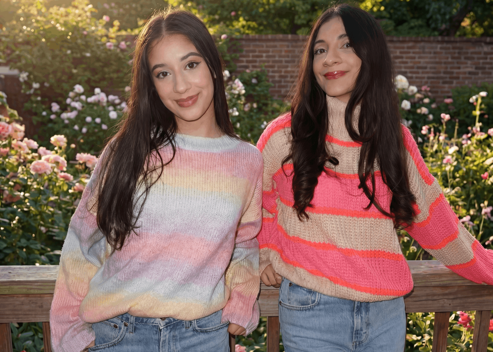 Two women wearing colorful striped sweaters standing outdoors with flowers in the background